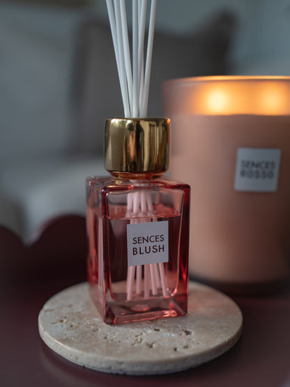 A Sences Blush reed diffuser bottle with a gold cap and white reeds, placed on a round stone coaster in front of a lit Senses Rosso candle.