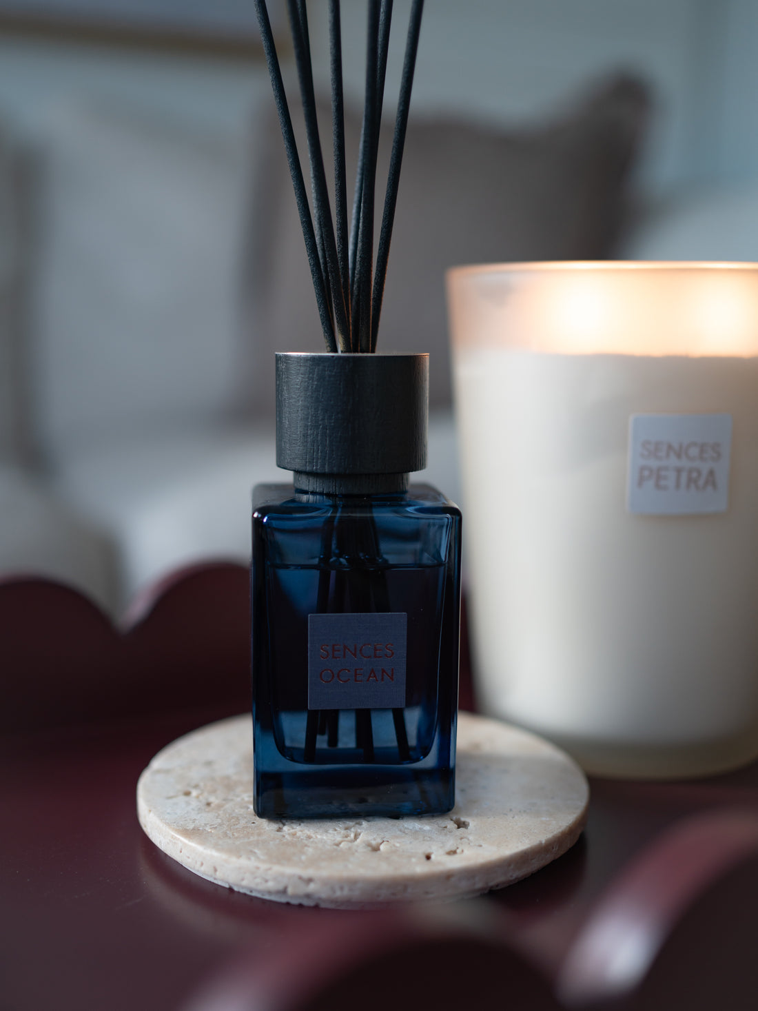 A dark blue glass small ocean reed diffuser bottle with black reeds and a black cap, placed on a round stone coaster next to a lit cream-coloured candle labeled &