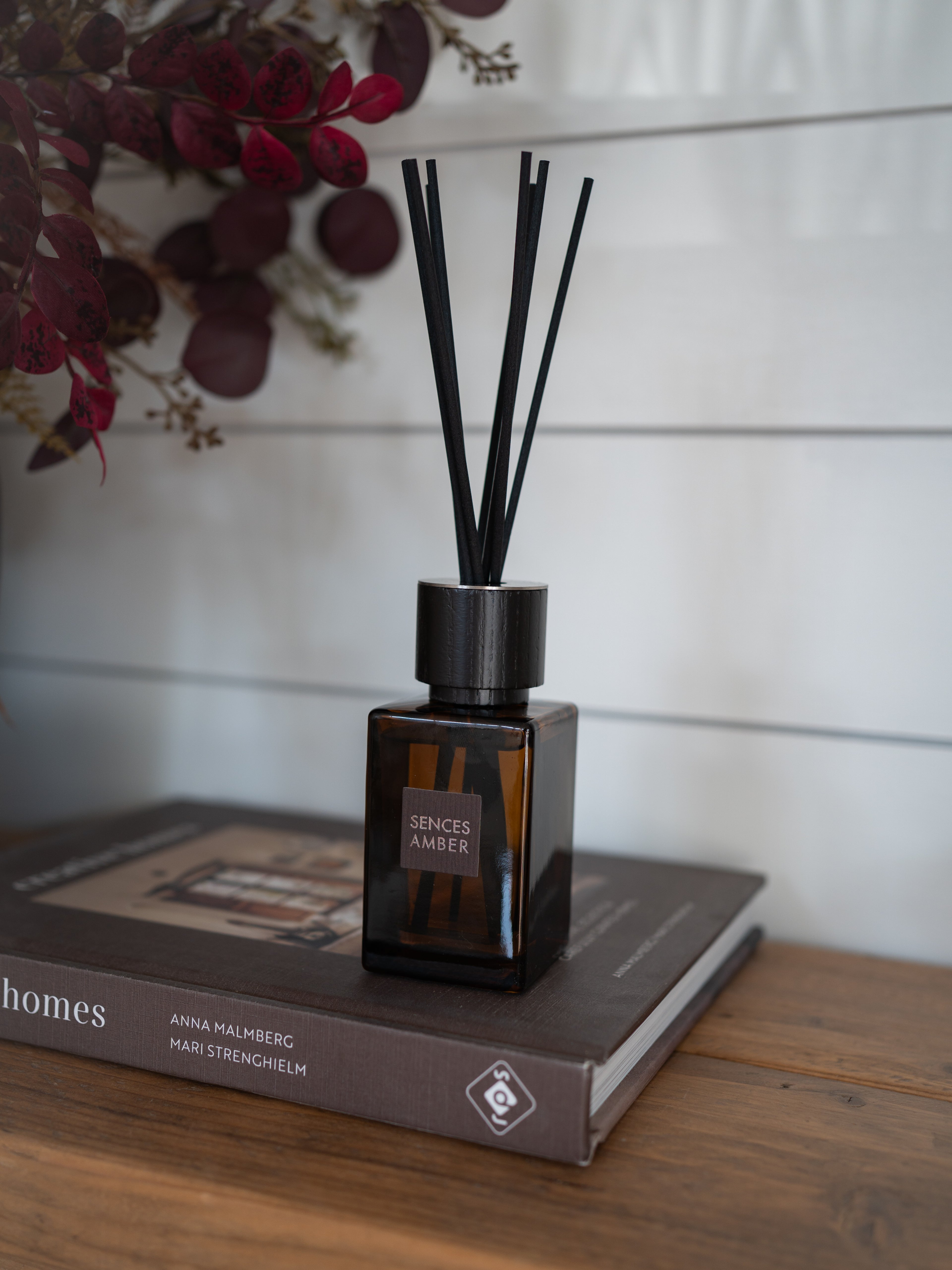 Amber glass reed diffuser bottle with black reeds, resting on a brown book on a wooden table against a white paneled wall.