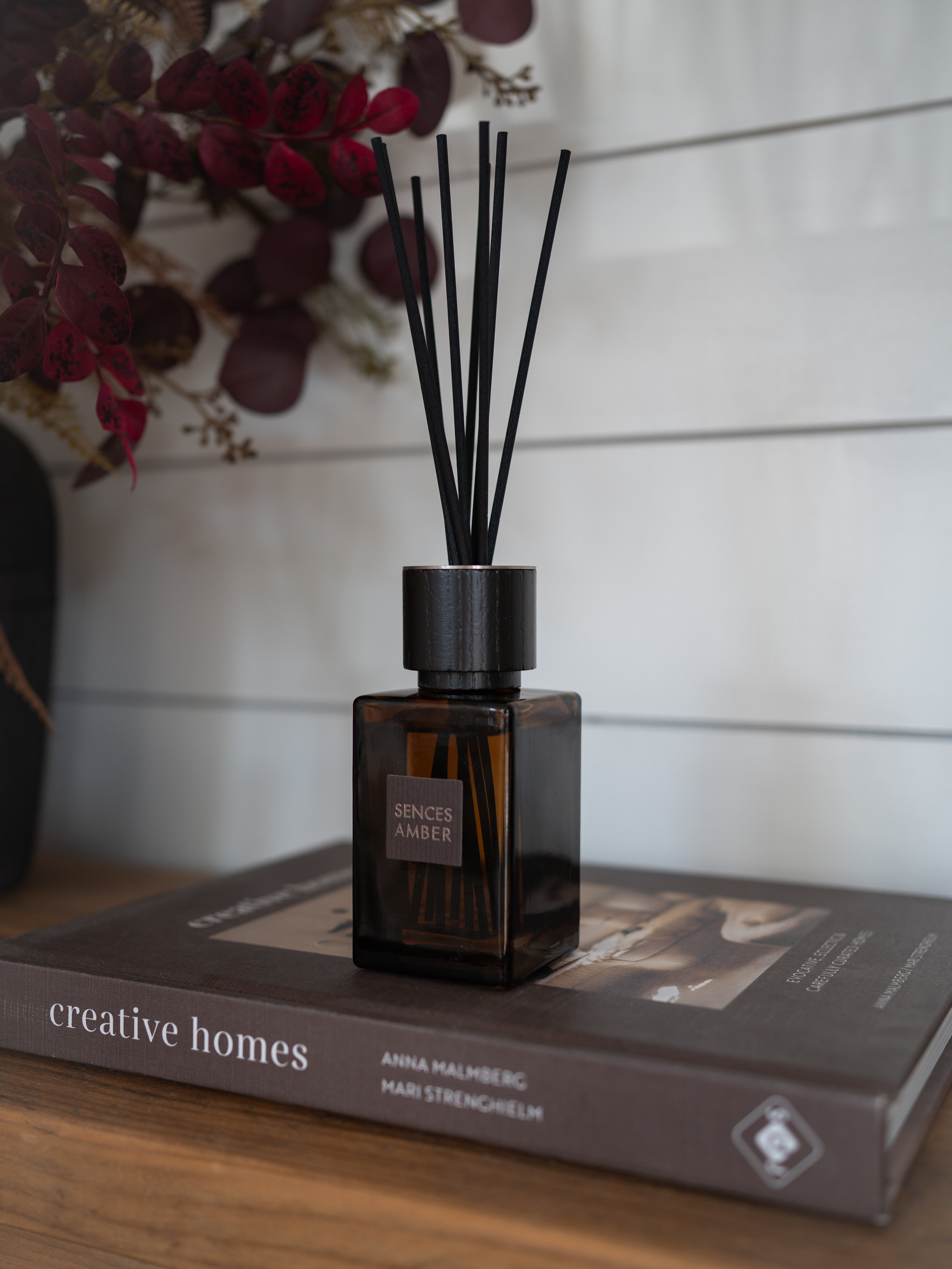 An sences amber alang alang glass reed diffuser bottle with black reeds sits on a brown book on a wooden surface, against a white shiplap wall with dried burgundy foliage in the background.