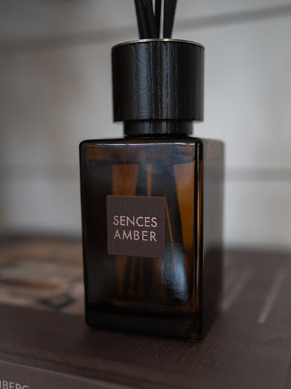 A Sences Amber reed diffuser bottle made of dark amber glass, featuring a black cylindrical cap with diffuser sticks and a brown label on the front.