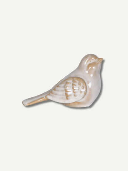 A small ceramic bird figurine in a glossy off-white glaze with warm tan and light brown painted details, shown on a plain light beige background.