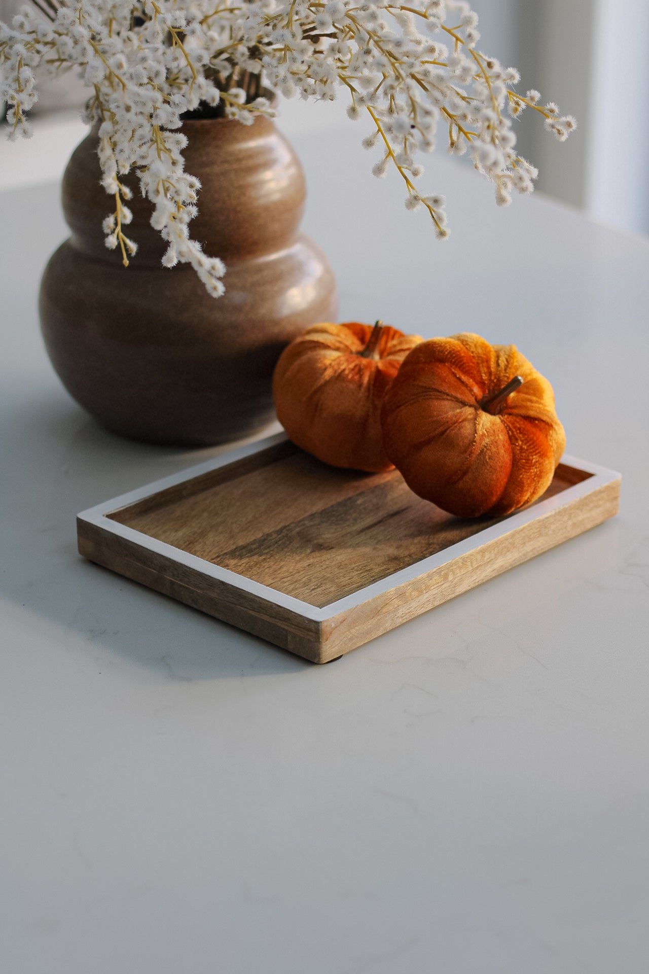 A small mango wood tray with white edge detailing, placed on a table with a vase of flowers and two orange pumpkins on top.