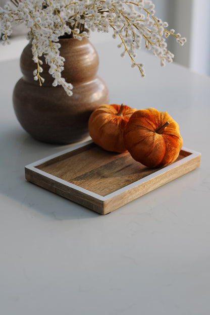 A small mango wood tray with white edge detailing, placed on a table with a vase of flowers and two orange pumpkins on top.