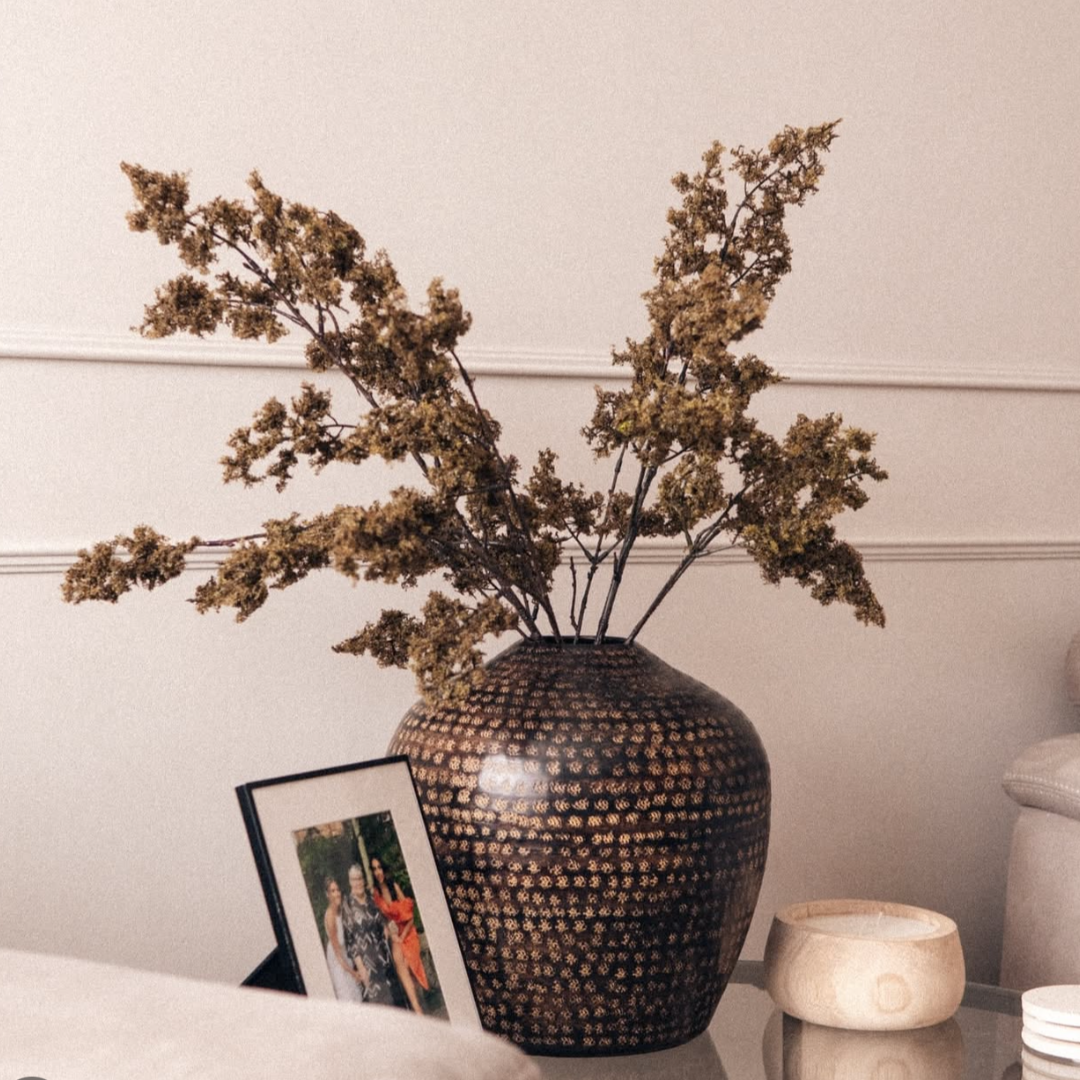 Hammered brown etched rounded metal vase with faux moss stems on side table in living room