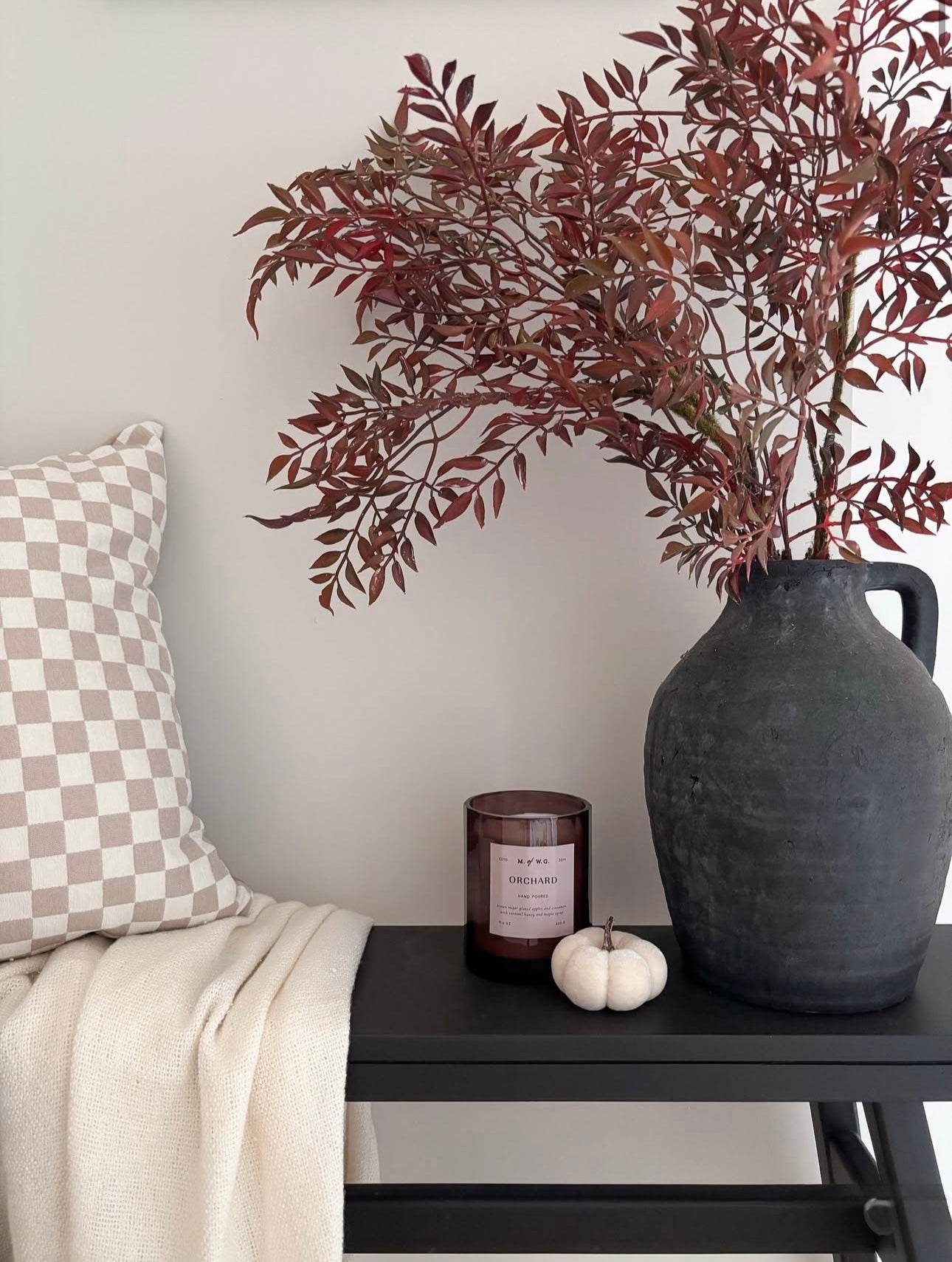 Decorative setup with a vase, candle, and checkered pillow on a bench against a plain wall.