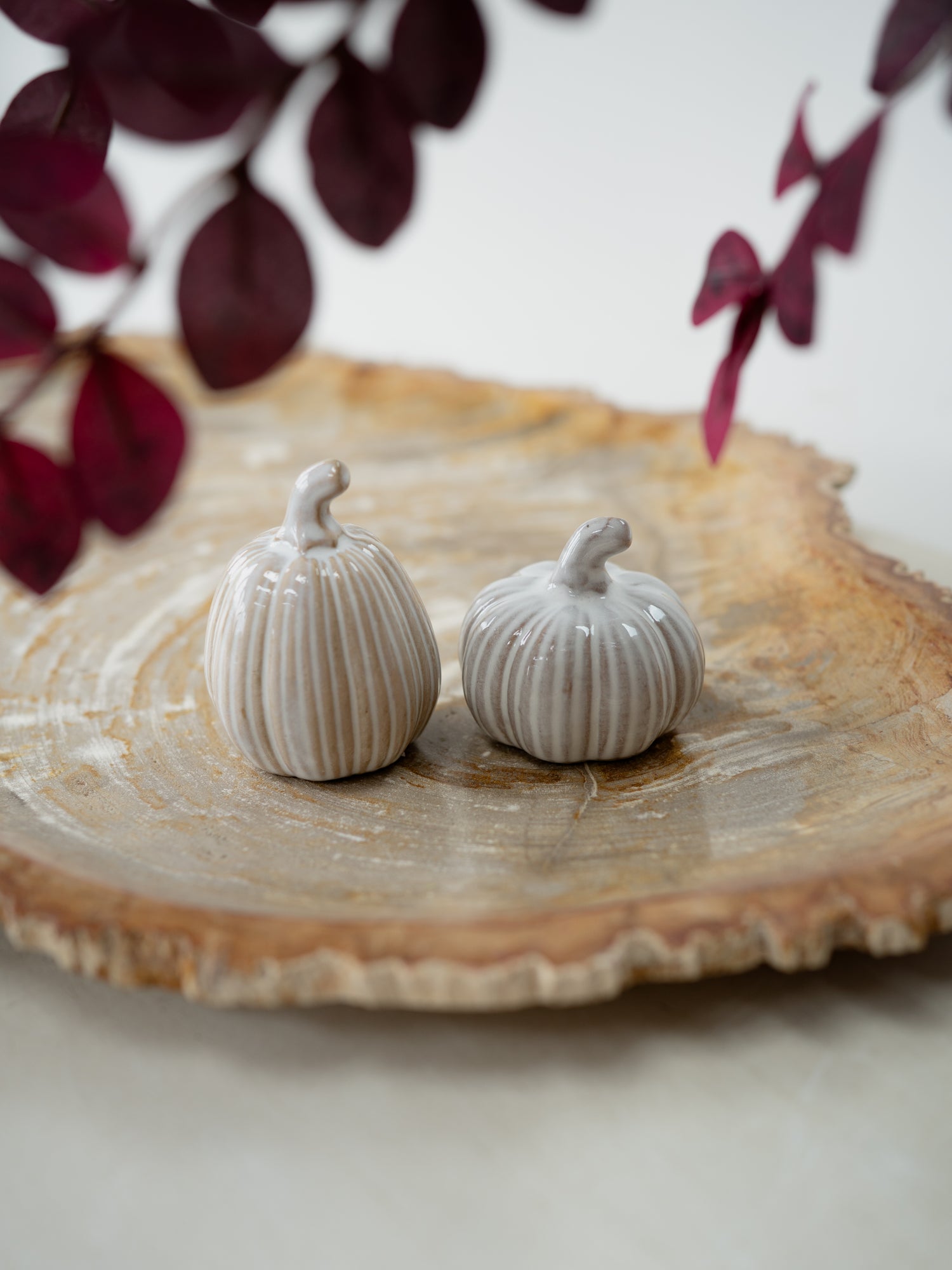 Two small ribbed ceramic pumpkins in a light warm gray or off-white glaze, displayed on a rustic round wood slice platter with blurred dark burgundy leaves in the background.