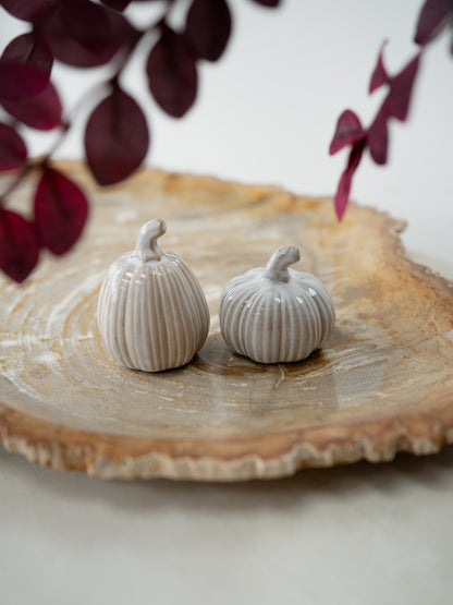 Two small ribbed ceramic pumpkins in a light warm gray or off-white glaze, displayed on a rustic round wood slice platter with blurred dark burgundy leaves in the background.