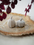 Two small ribbed ceramic pumpkins in a light warm gray or off-white glaze, displayed on a rustic round wood slice platter with blurred dark burgundy leaves in the background.