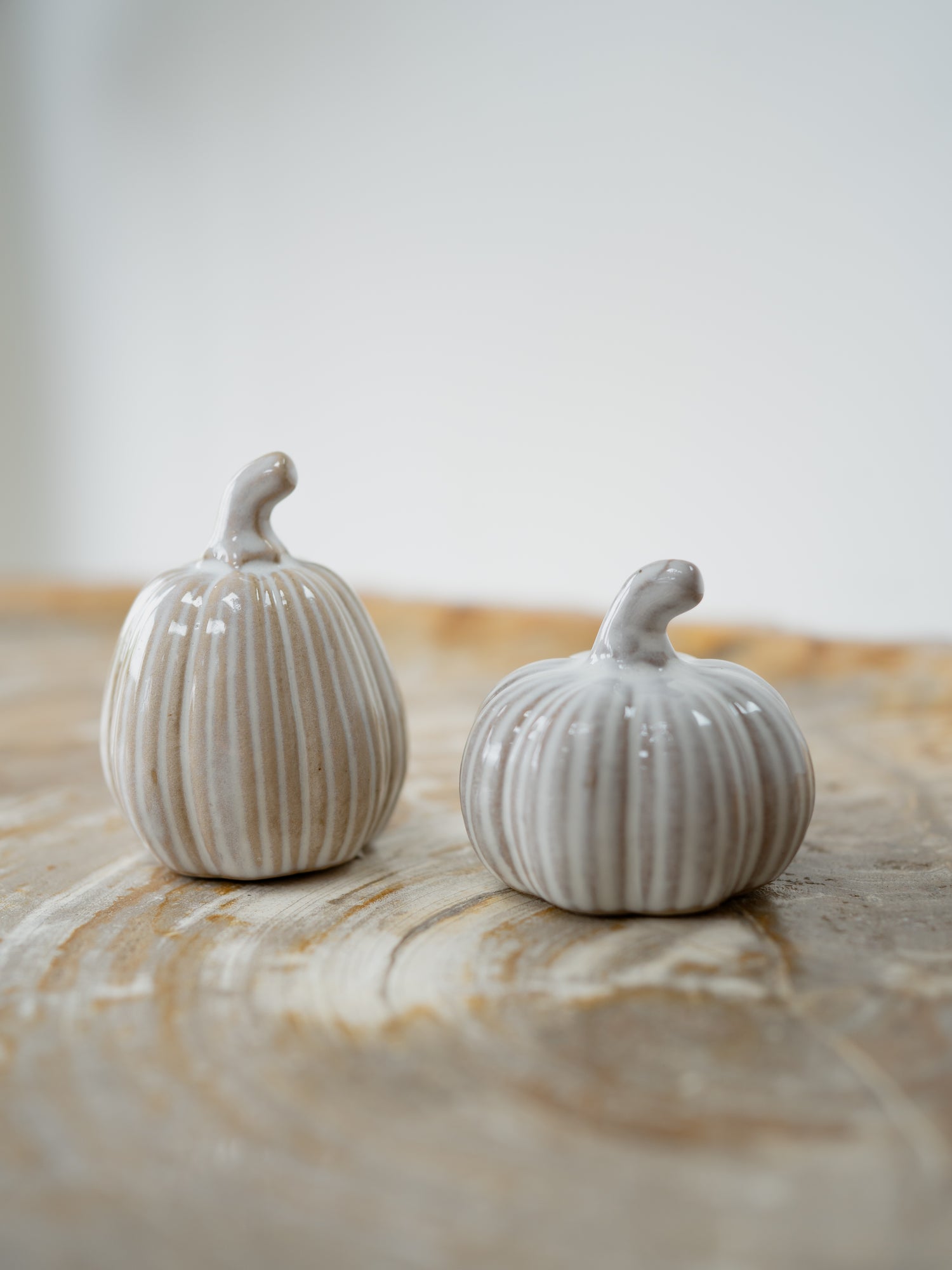 Two small ribbed ceramic pumpkins in a warm off-white glaze, displayed on a light wood surface against a soft neutral background.