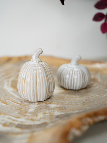 Two small ribbed decorative pumpkins in an off-white glazed ceramic finish, displayed on a round wooden slice tray.