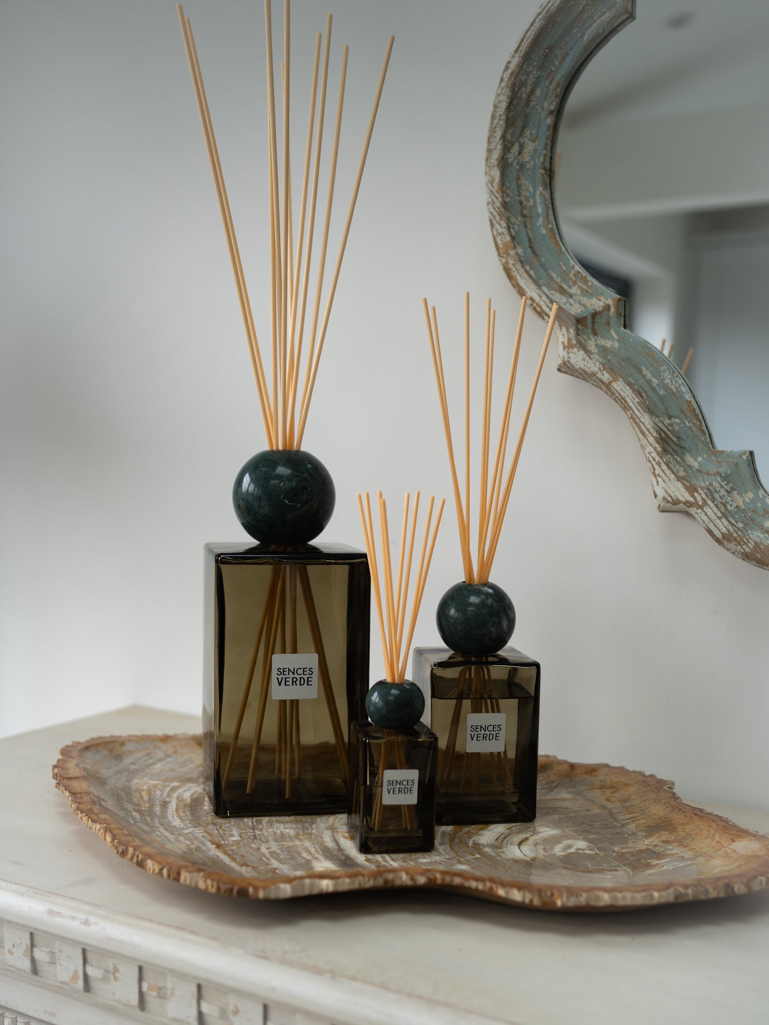 Three amber green reed diffusers with dark green spherical marble tops and natural reed sticks, arranged on a decorative stone tray against a light wall with a rustic mirror partially visible.