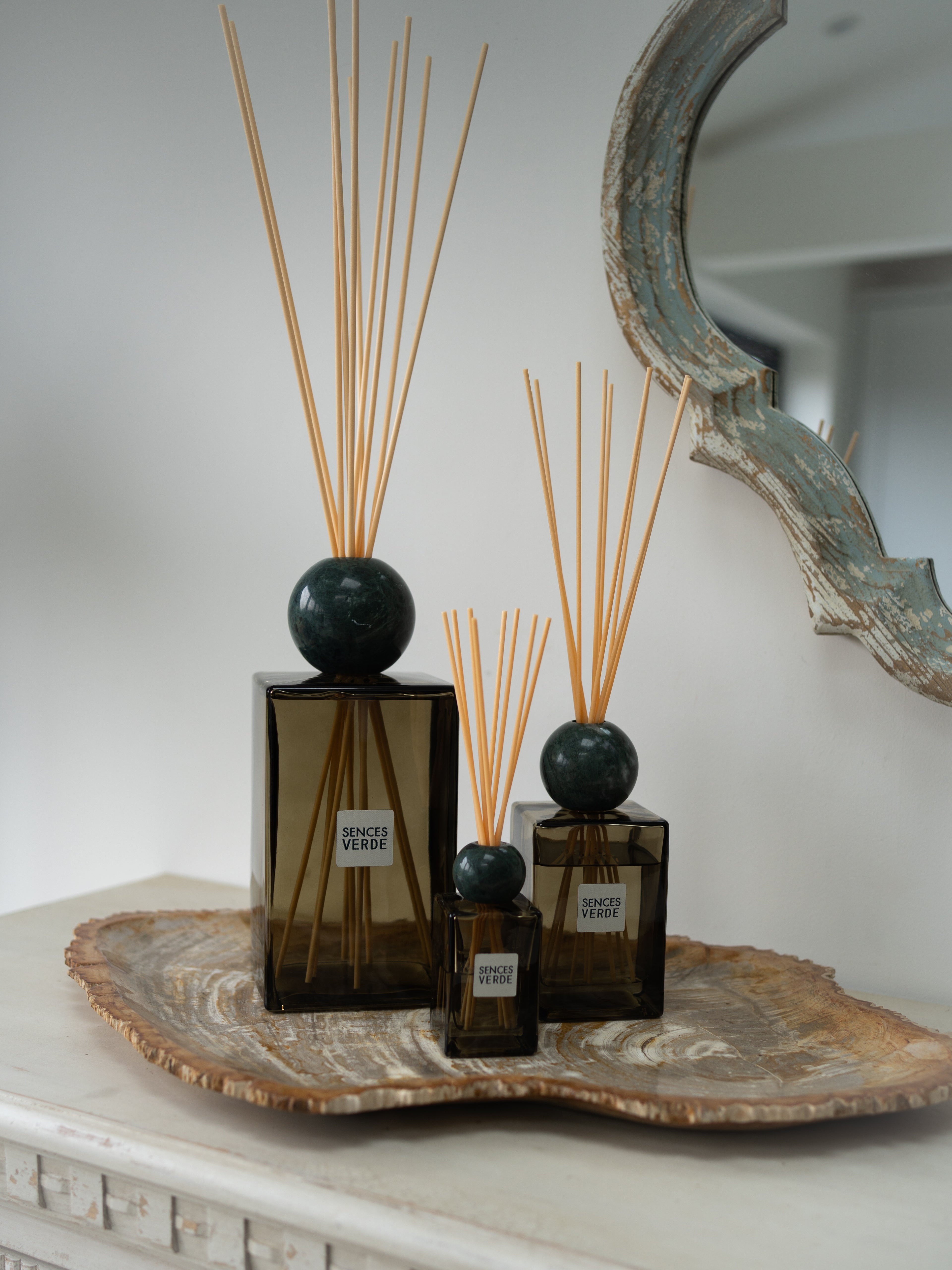 Three amber green reed diffusers with dark green spherical marble tops and natural reed sticks, arranged on a decorative stone tray against a light wall with a rustic mirror partially visible.