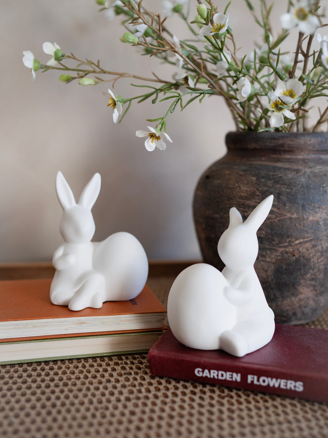 Two white ceramic rabbit sculptures on books with a vase of flowers in the background.