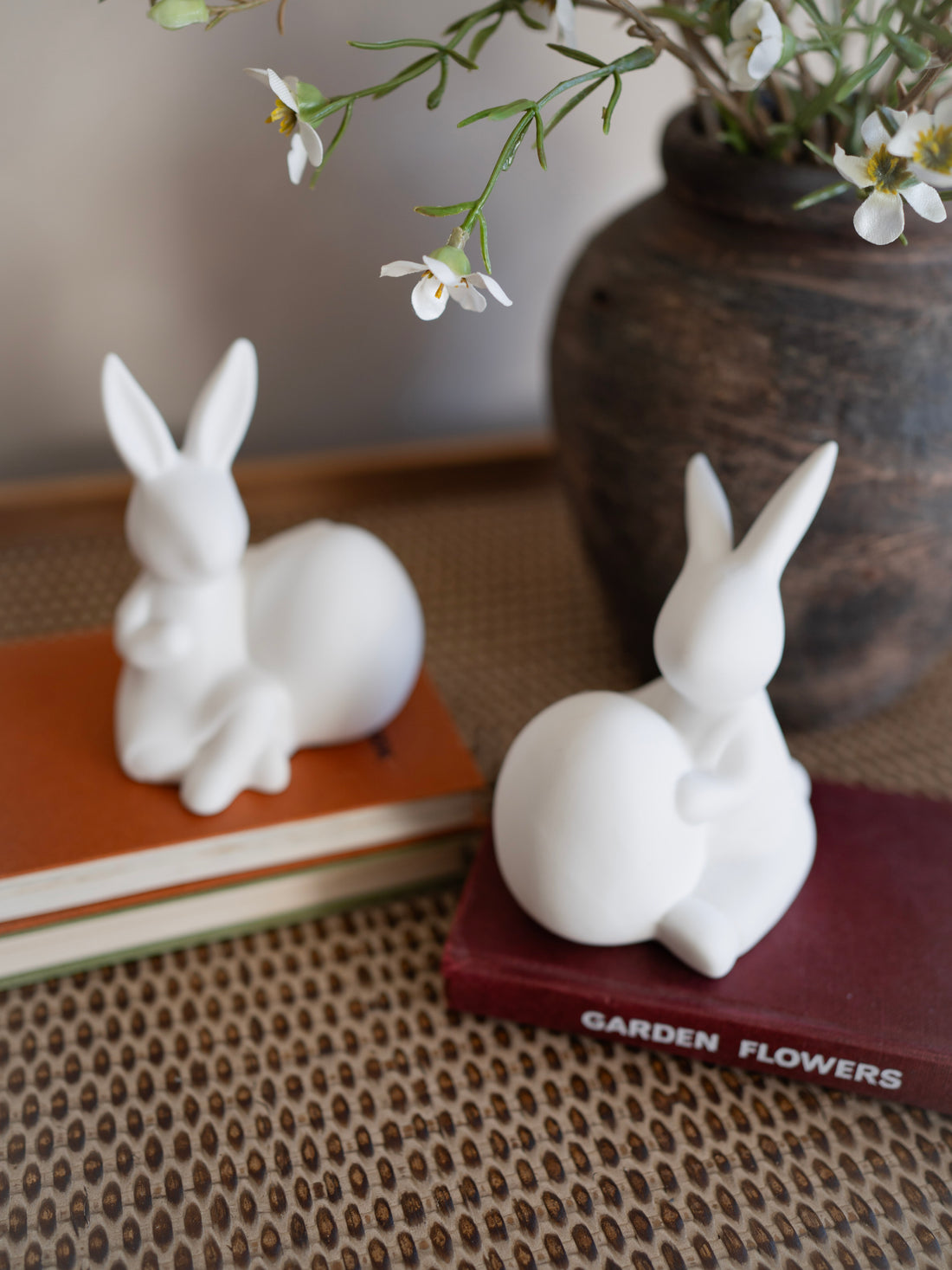 Two white rabbit figurines on books with a vase of flowers in the background.
