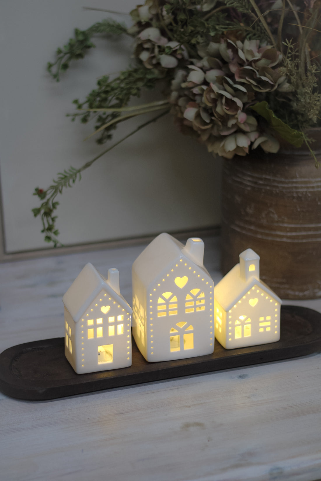 White ceramic houses with cut-out hearts and dots design, illuminated from the inside by LED lights, placed on a wooden surface.