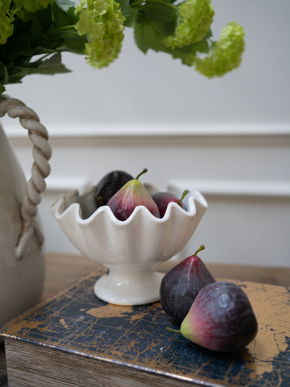 White decorative bowl with faux figs on a textured surface with a floral arrangement in the background. Beautiful home decor and styling