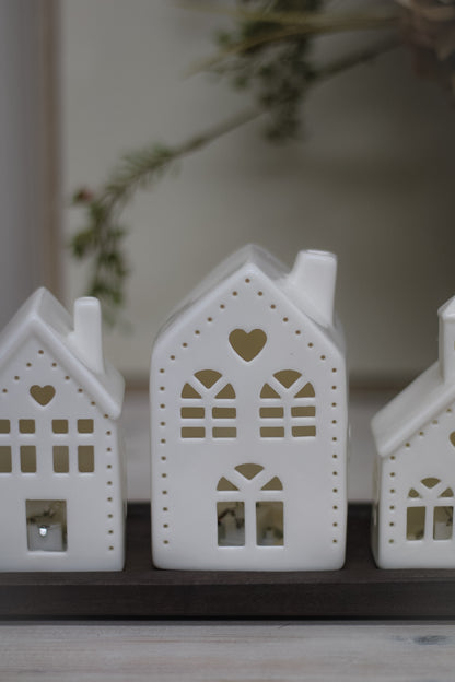 White ceramic houses with cut-out hearts and dots design, illuminated from the inside by LED lights, placed on a wooden surface.